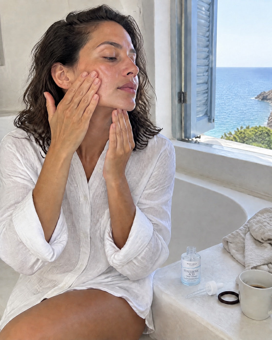 A woman by an open window smoothing serum across her cheek, the open Hyaluronic Acid bottle and its dropper beside her